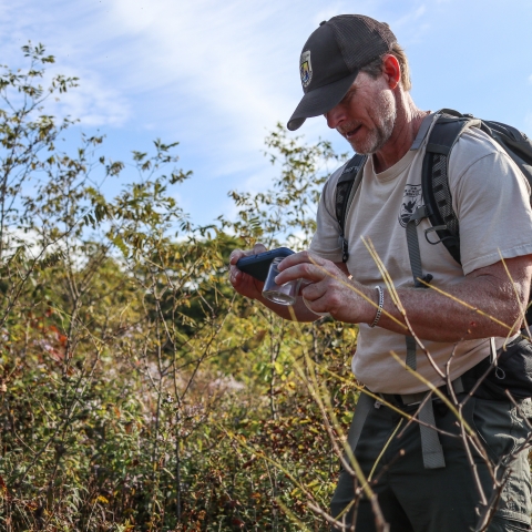 Male biologist stands in a field with a phone and glass jar in his hands