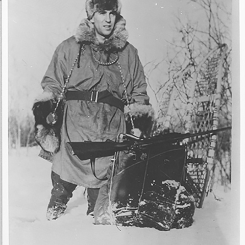 Clarence J. Rhode appears warmly dressed and holds a rifle while standing in the snow in Alaska. 