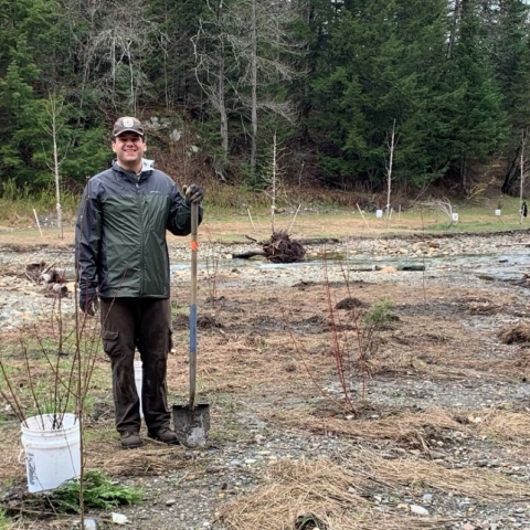 Dave Rojek, shovel in hand, standing near freshly planted trees at a riparian project site.