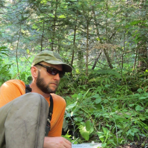 Fish Biologist, Brian Davis, collecting samples in the field. Brian is wearing an orange shirt, tan pants, tan hart and black sunglasses. He is reclining on the ground in a temperate rainforest setting with sampling equipment in front of him. 