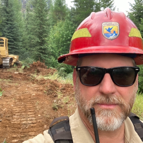 A man in sunglasses and a red protective hard hat standing in an opening with a bulldozer in the distance