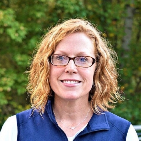 a blonde woman wearing glasses, a white shirt and blue vest smiling in front of a forest background.