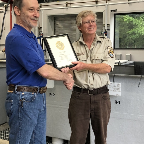 Man in brown uniform receives an award plaque from another man in blue while standing next to gray fish tanks.