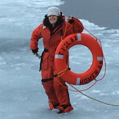 Woman stands in icy surf wearing orange float coat and holding up orange life ring.