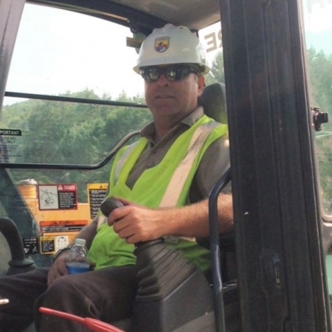 Phil Herzig with USFWS hard hat and work vest inside cab of excavator. 
