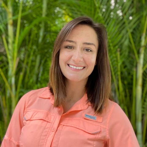 Jessica Sutt smiles, standing in front of topical plants, for a profile photo