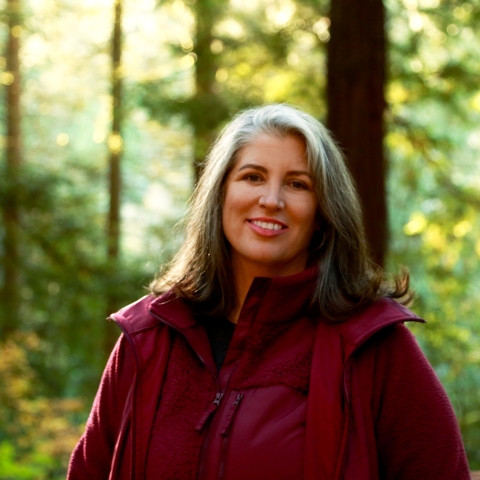 Photo of woman in a burgundy colored coat smiling, posing for photo in front of fir trees with sun shining through the trees 
