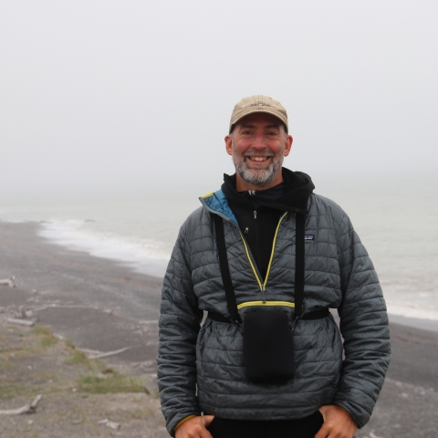 Man standing along Alaska coastline with binoculars around neck