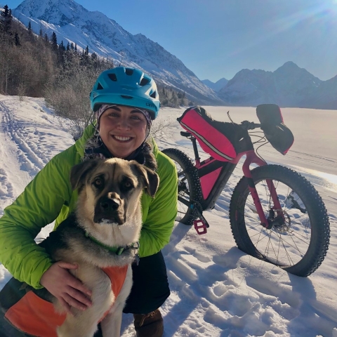 A picture of a woman next to a dog with a bicycle and snowy mountain landscape in the background