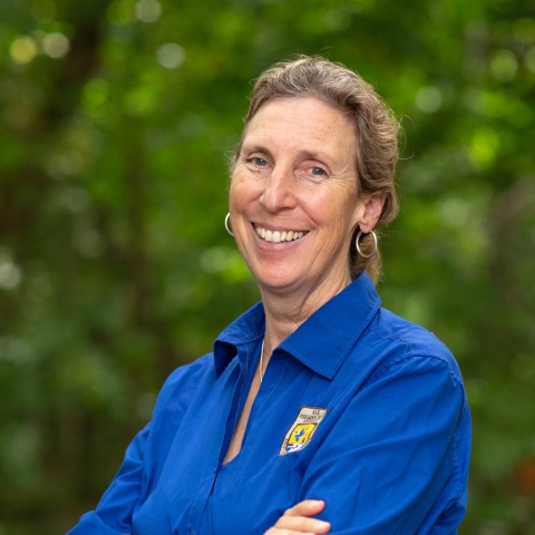 Former Director Martha Williams stands outdoors, smiling, with arms folded, wearing a blue shirt with FWS logo. Behind her is a wooded area.