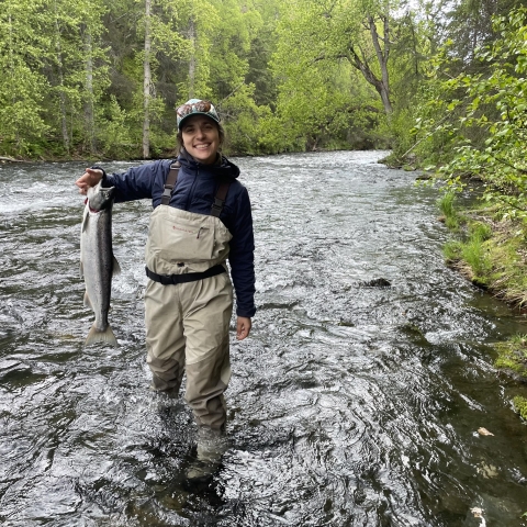 A person wearing wader overalls stands mid-calf in a river. They hold a fish in their hand with the other at their side while they smile.