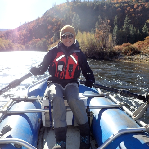 A person in a life vest sits in an inflatable boat holding oars. The boat is is the middle of river surrounded by forests.