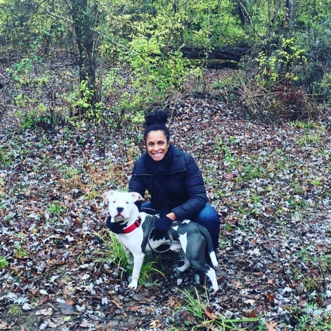 Nancy kneels down in a wooded area with dog.