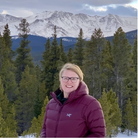 Nicole smiles in a purple puffer coat amid pines and a mountain landscape