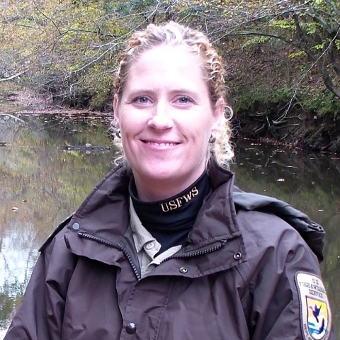 Woman smiling and wearing a brown FWS uniform. A stream and vegetation serve as backdrop.