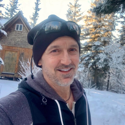 man in winter gear stands in front of a snowy cabin