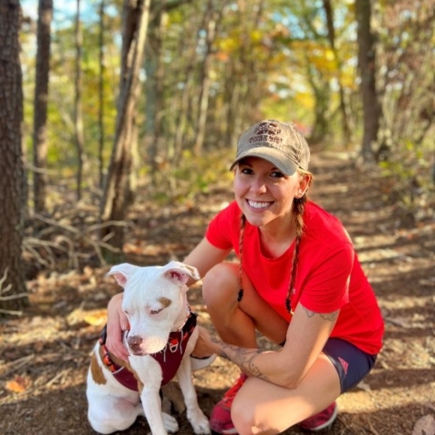 Shannon kneels and smiles with her arms wrapped around a white and brown spotted pitbull. She wears black running shorts, red running shoes, a red t-shirt, and a tan baseball cap