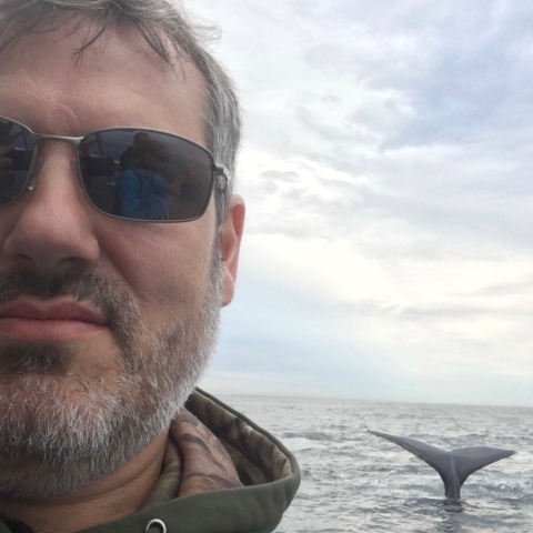 a selfie of a man in a boat with a whale's tail emerging from the water behind him