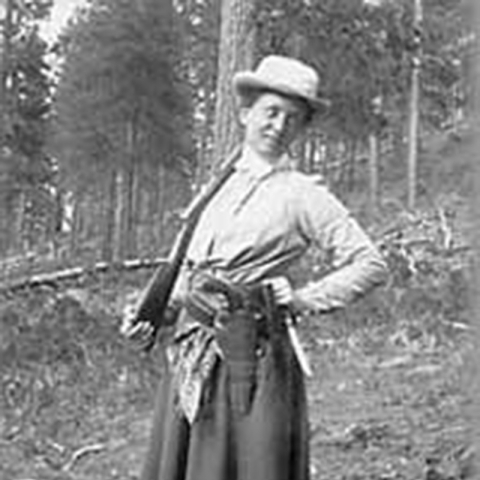 smiling woman standing holding rifle on shoulder with wood in background