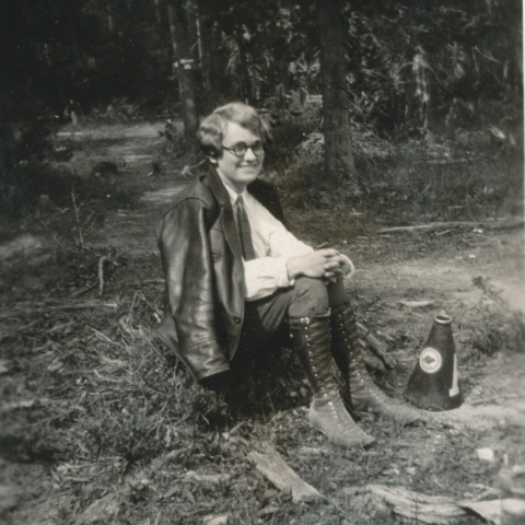 woman with short hair and glasses sitting on ground smiling near wooded area