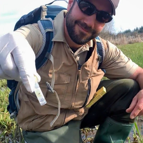 A smiling man with a black beard and dark sunglasses, wearing waders and a Service hat, kneels in a wetland and holds out a vial toward the camera.