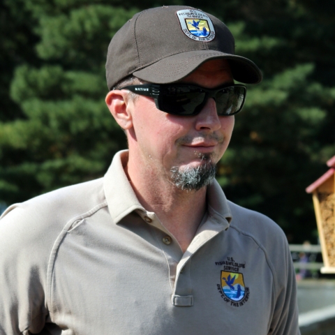 A man in Service short-sleeved polo shirt and ballcap, dark sunglasses, and goatees smiles slightly.