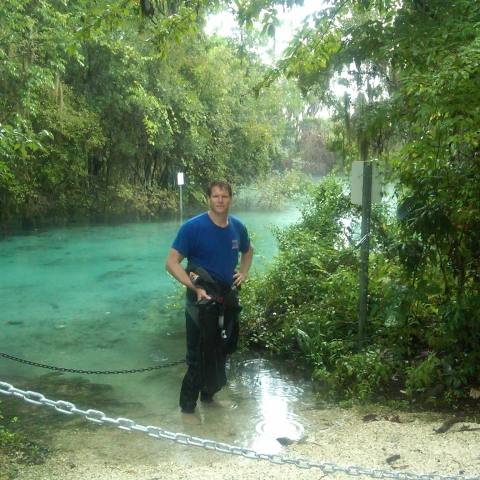 Man stands ankle-deep in blue water half-dressed in a wetsuit.