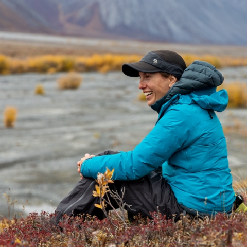 side view of woman sitting on riverbank tundra, with gravel, plants and base of mountain in background