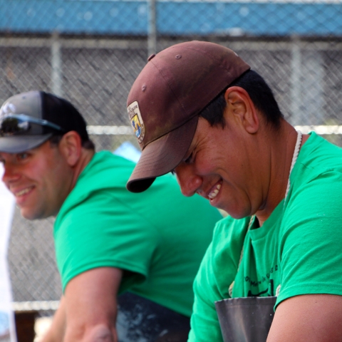A man in green t-shirt and Service ballcap grins while looking down at a task, with another man behind dressed in identical t-shirt and a non-Service ballcap also smiles while looking up.