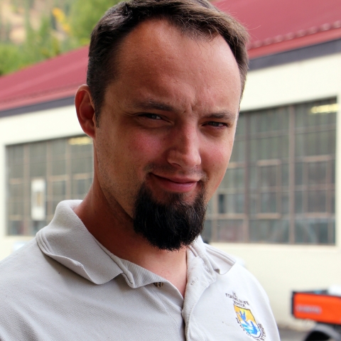 A man in Service polo shirt smiles into camera, with a red-roofed building behind him with multi-paned windows all along its length.