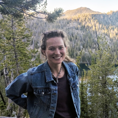 woman from waist up, in front of spruce forest and mountain top.