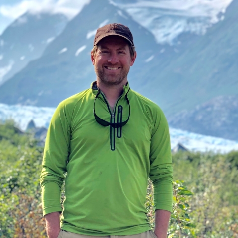 A man standing outside with a field, a glacier, and mountains in the background