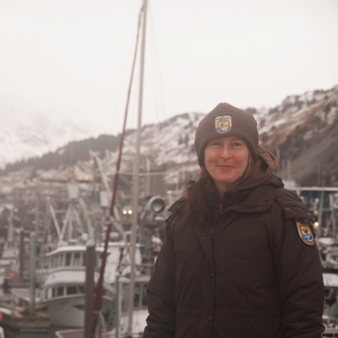 woman in usfws uniform stands in front of harbor with snowy mountains in the background