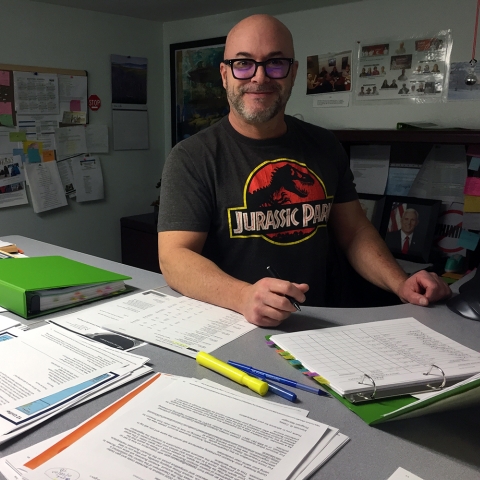 A man in glasses and a Jurassic Park t-shirt stands at a desk covered in short stacks of organized paperwork.