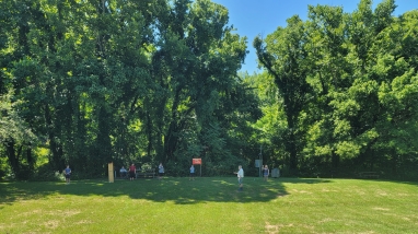 Short green grass field with a group of kids and trees behind a fly fishing instructor