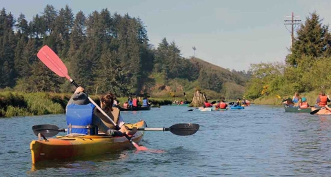More than a dozen people in kayaks and canoes move down a river banked by pine trees