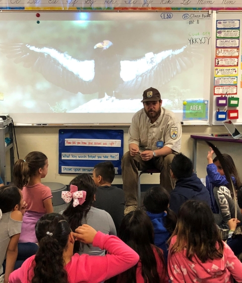 A Park Ranger sits infront of an projected image of a condor sunning on a rock perch, wings streched wide. In front sitting on the floor are about 10 3rd grade students listening.