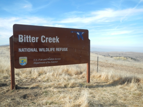 A brown sign reads Bitter Creek National Wildlife Refuge. The sign is large, about half the image, and is on a grassy hilltop overlooking a long view shed with blue sky and white wispy clouds.