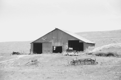 a black and white photo showing an old tin barn on a grassy slope with old farm equipment strewn around.