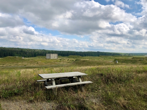 A wooden picnic table sits overlooking a field with an abandoned military structure in the distance. Huge billowy clouds fill the sky above. 