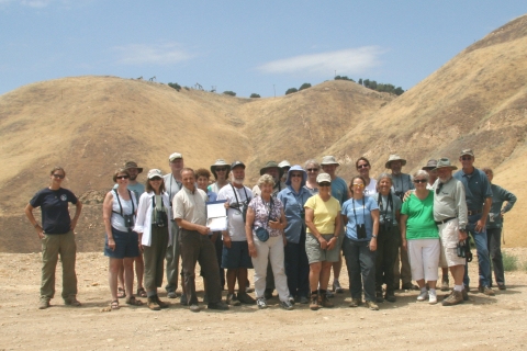 A group of about 20 people in various clothes and hats stand for a photo in front of golden rolling hills beneath a blue sky.