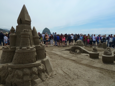 A crowd observes a sandcastle-building contest with Haystack Rock visible in the distance