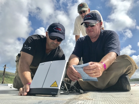 Two US Fish and Wildlife Service personnel sit and kneel around a device on a roof in order to give phone and internet service to the Vieques National Wildlife Refuge