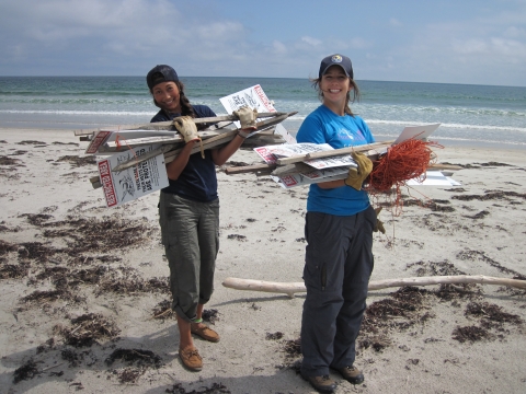 Two interns collect signs along the beach and smile at the camera