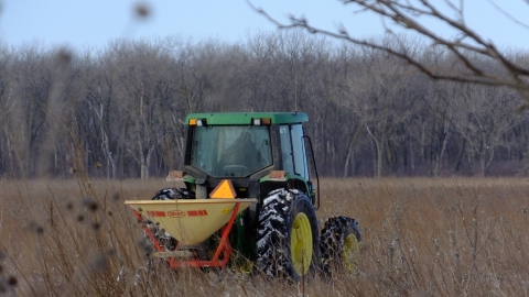 A tractor with a seed spreader on the back driving through a prairie field spreading seed. 