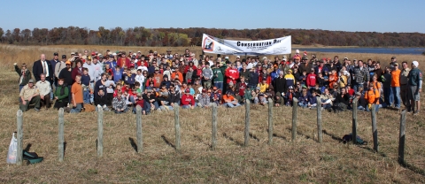 A large group of people stand in front of a wetland with a sign that reads, "conservation day"