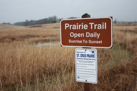 A prairie with a sign introducing visitors to the regulations, open daily sunrise to sunset, Waterfowl Production Area, Managed by the U.S. Fish and Wildlife Service, Purchased with Duck Stamp Dollars, Help protect wildlife follow these regulations, open to foot access only, dogs must be on a leash, except while hunting, nontoxic shot is required for ll hunting, blinds and tree stands must be removed daily, additional regulations apply, for more information contact: St. Croix Wetland Management District