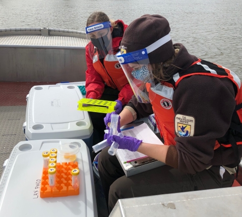 Two people in brown uniforms and masks sit in a boat with a tray of vials, with the river and forest in the background