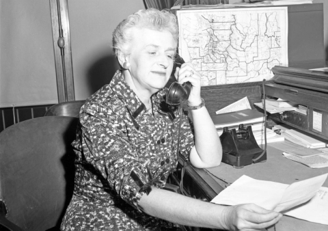 A black and white photograph depicting an older woman talking on the phone while looking at a piece of paper in an office with maps. She is smiling slightly.
