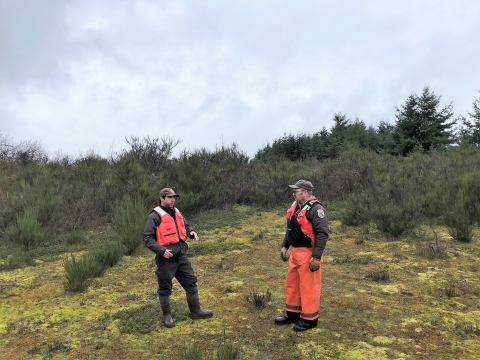 Two men in brown uniforms with orange safety gear stand on marshy ground with green vegetation behind them.